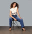 Woman seated wearing mid-rise blue jeans and a sleeveless white top with ruffle details, paired with beige heeled sandals, posing against a neutral background.
