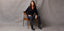 Woman looking to the side wearing a black button-down top paired with black frayed hem jeans, sitting on a wooden chair against a gray background.
