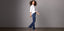 Woman in a white shirt and blue bootcut jeans walking confidently in heels against a plain gray background