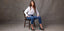 Woman wearing a white button-down top paired with blue cropped petite jeans, sitting on a wooden chair against a grey background.