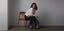 Woman wearing wide jeans with a stylish white sweater and black heels, posing elegantly on a wooden chair against a minimalist gray backdrop.