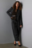 Fashionable woman wearing a dark tailored blazer and relaxed barrel leg trousers, paired with a belted knit sweater and black heels, leaning casually against a gray wall.